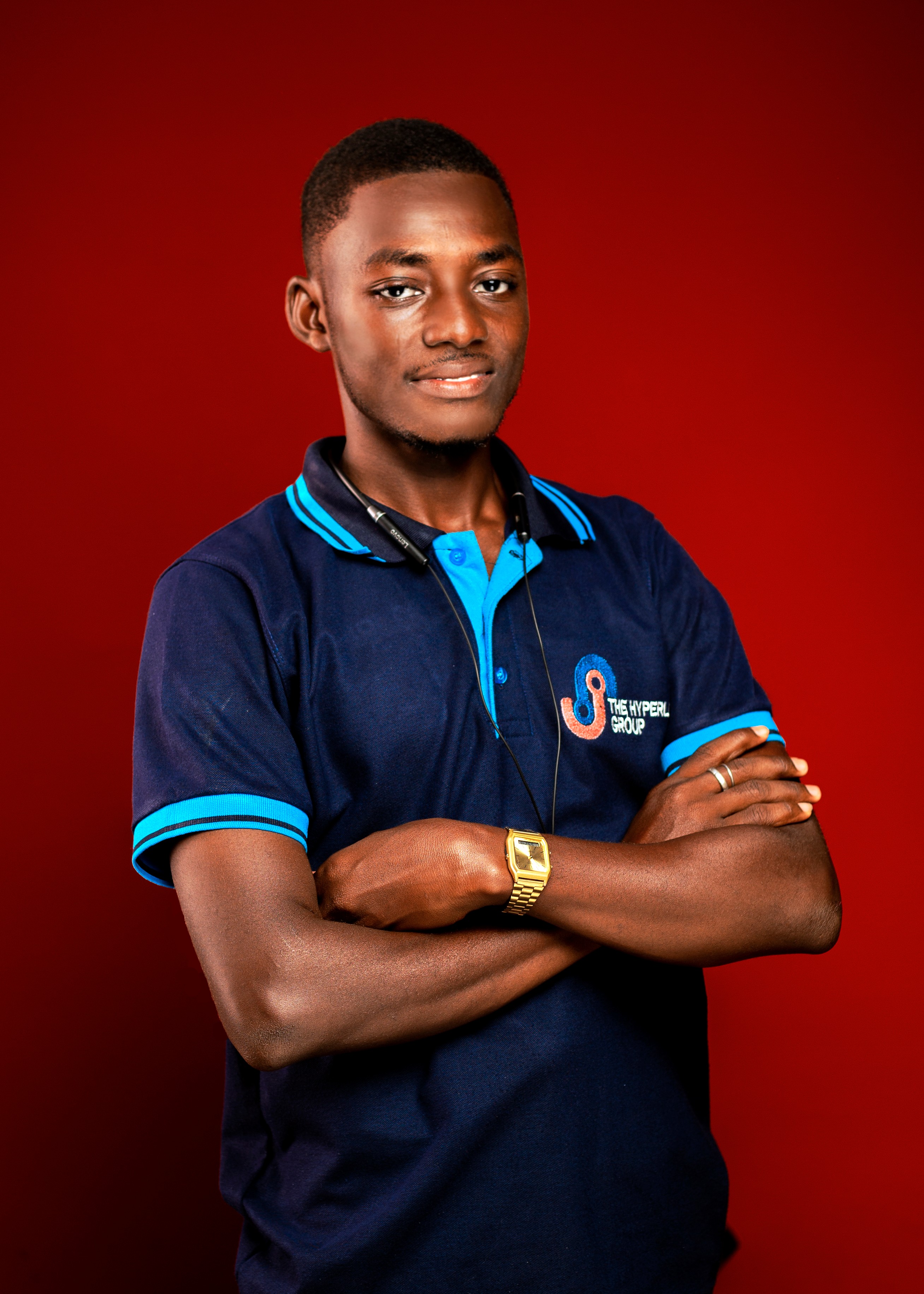 Young man with arms crossed, wearing a navy blue polo shirt with blue accents, standing against a red background.