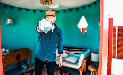Person wearing a cap and denim jacket pointing at the camera in a room with teal walls and a bed.