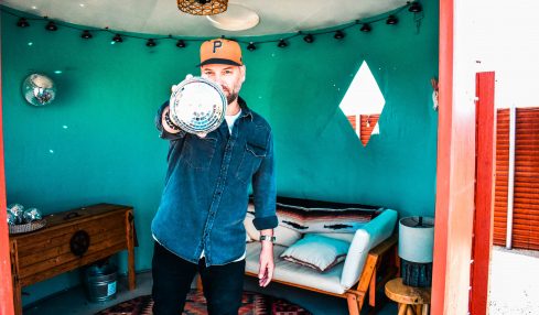 Person wearing a cap and denim jacket pointing at the camera in a room with teal walls and a bed.