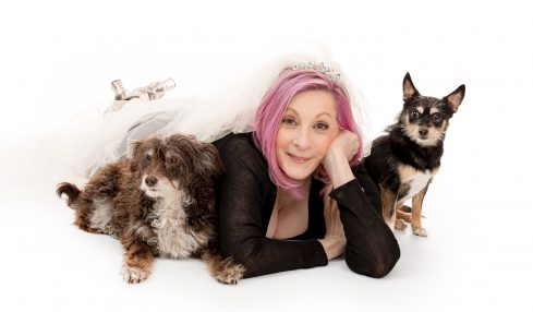 Woman with pink hair lying on her stomach with two dogs, one fluffy and one small, on a white background.