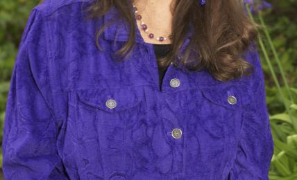 Woman with long brown hair smiling outdoors, wearing a purple jacket and jewelry, surrounded by greenery and purple flowers.