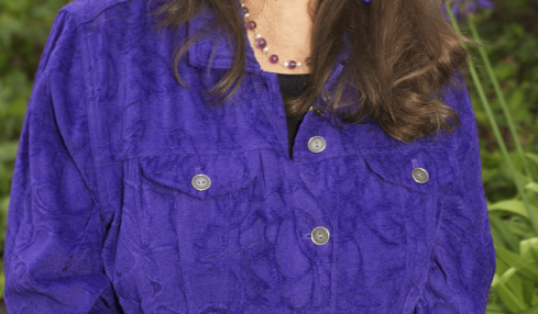 Woman with long brown hair smiling outdoors, wearing a purple jacket and jewelry, surrounded by greenery and purple flowers.