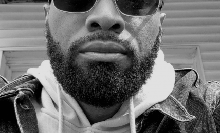 Man with sunglasses and beard wearing hoodie and denim jacket, looking at camera, outdoor setting, black and white.