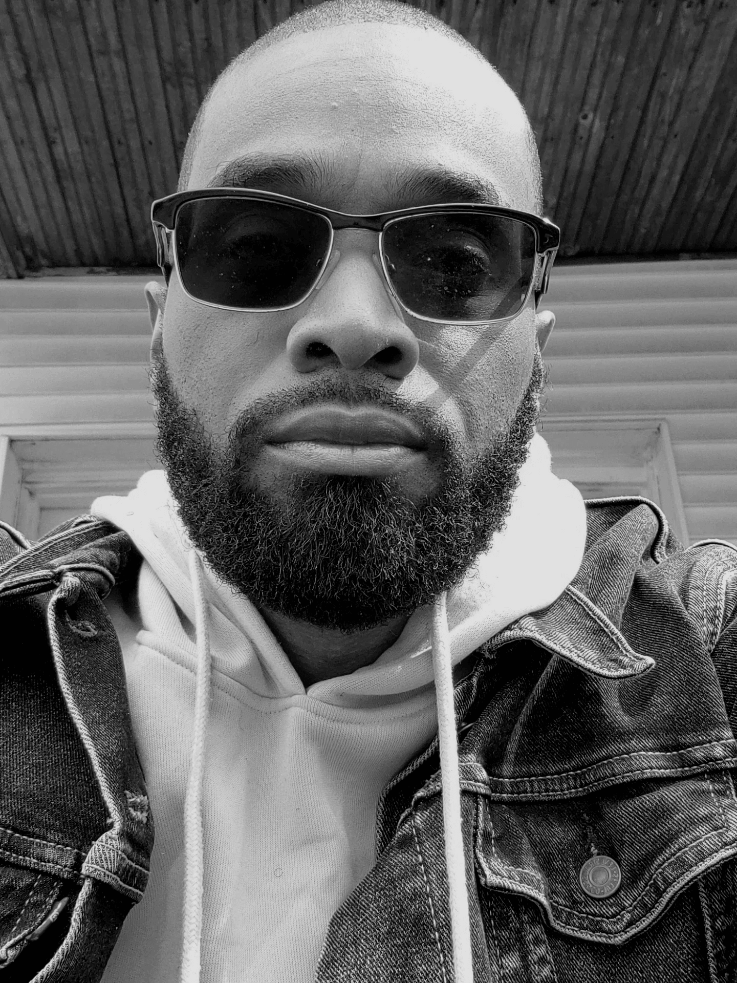 Man with sunglasses and beard wearing hoodie and denim jacket, looking at camera, outdoor setting, black and white.