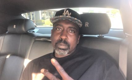 Man with a beard and mustache wearing a black cap and black shirt, sitting in a car, making a peace sign.