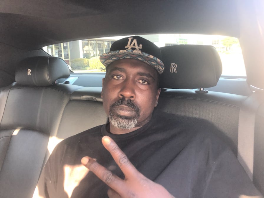 Man with a beard and mustache wearing a black cap and black shirt, sitting in a car, making a peace sign.