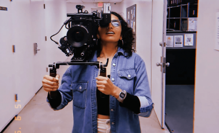 Person holding a camera in a hallway, wearing glasses and a denim jacket, taking a selfie.