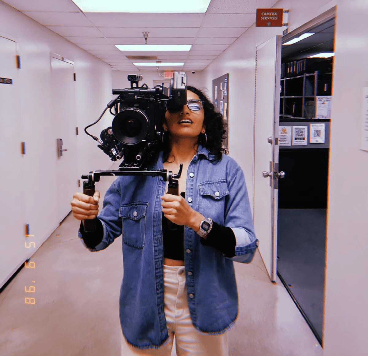 Person holding a camera in a hallway, wearing glasses and a denim jacket, taking a selfie.