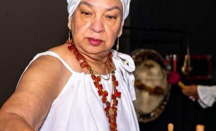 Older woman wearing a white headwrap and dress, holding a wooden mallet, with a large drum in front of her.