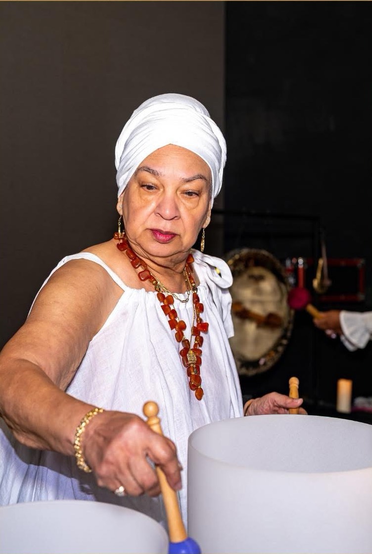 Older woman wearing a white headwrap and dress, holding a wooden mallet, with a large drum in front of her.