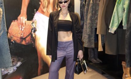 Woman in black crop top, striped pants, black jacket, sunglasses, holding a handbag, standing in clothing store.