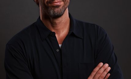 Man with dark hair and beard, wearing a black shirt, crossing arms, looking at camera, neutral expression.