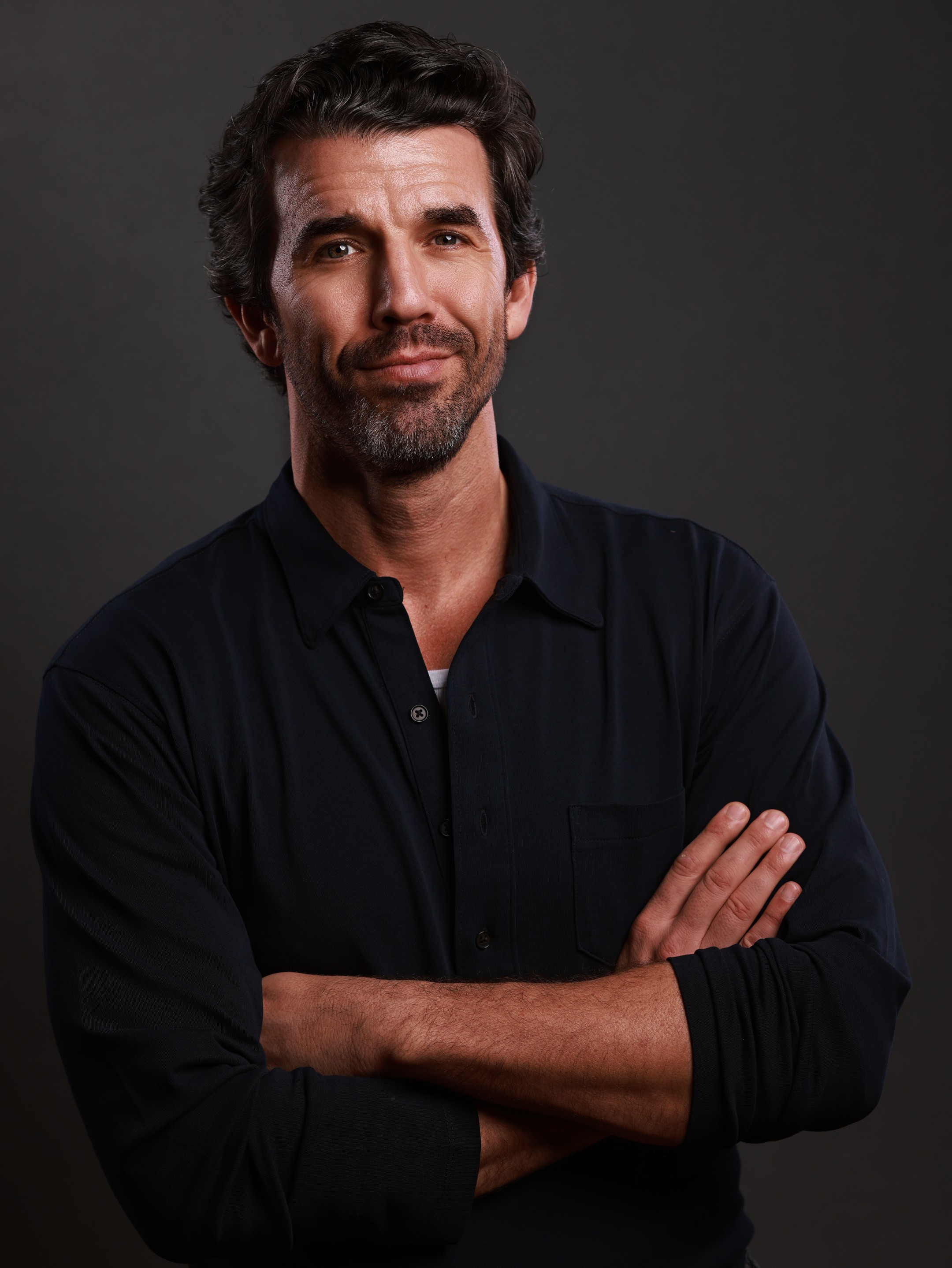 Man with dark hair and beard, wearing a black shirt, crossing arms, looking at camera, neutral expression.