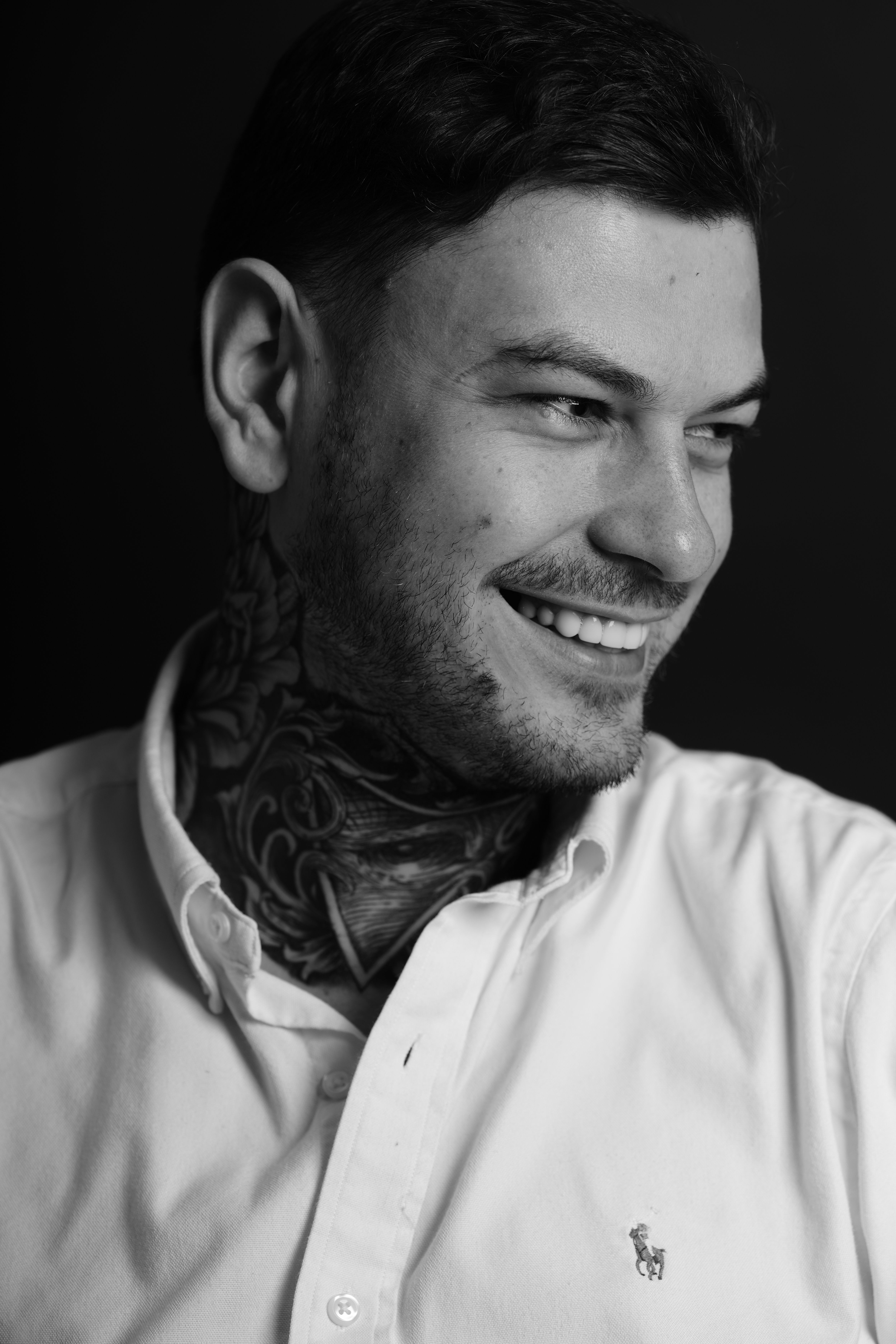 A man with dark hair and a beard smiling, wearing a light-colored shirt, facing right, against a dark background.