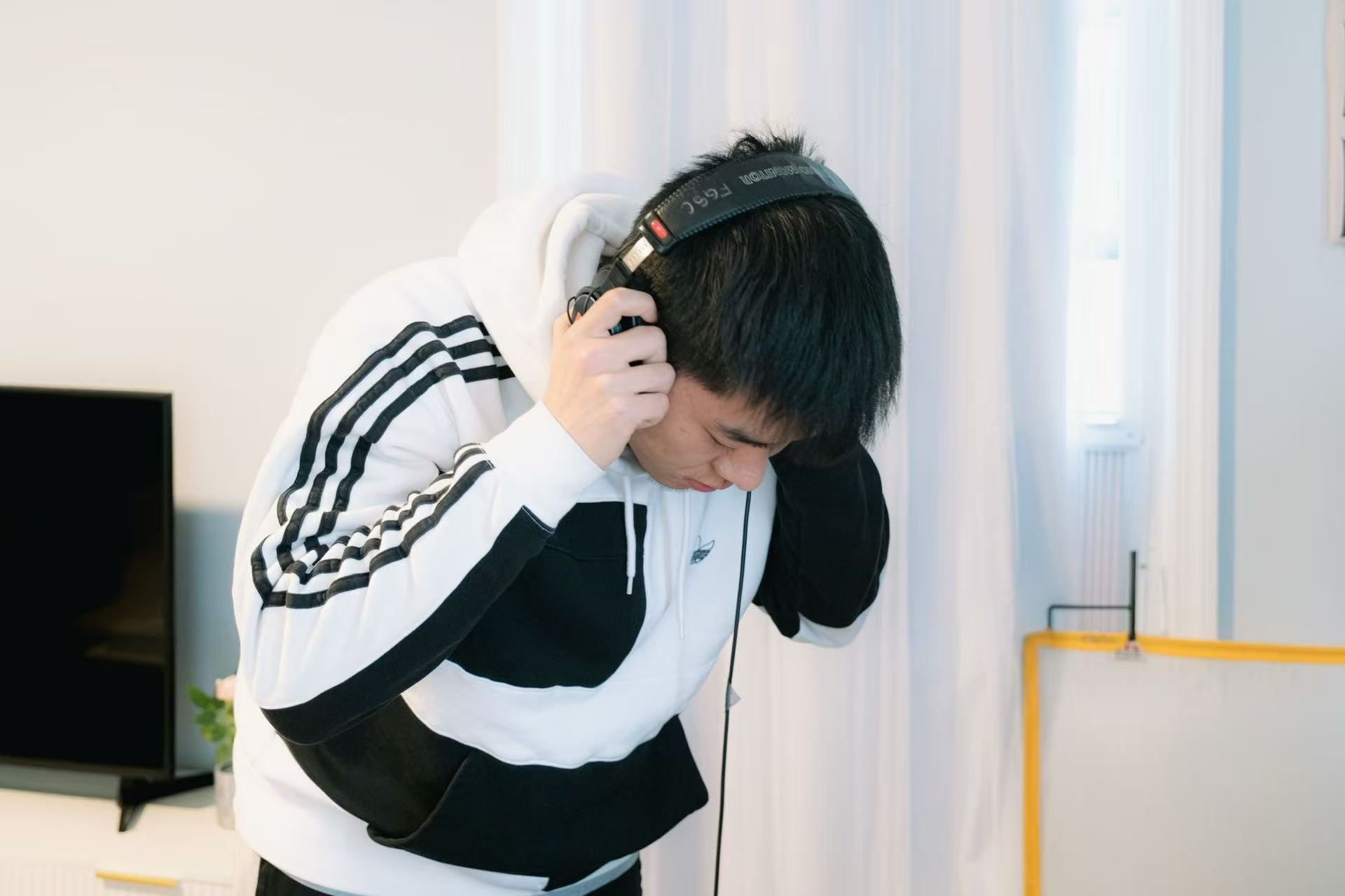 Person wearing headphones, leaning forward, in a bright room with curtains and a yellow object in the background.
