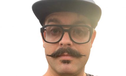 Person with glasses, mustache, wearing a cap, and a dark shirt, looking at the camera against a white background.