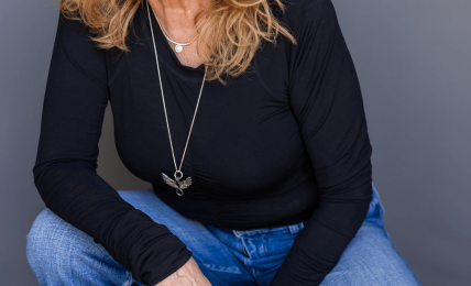 Woman with curly red hair smiling, wearing a black top and blue jeans, crouching against a gray background.