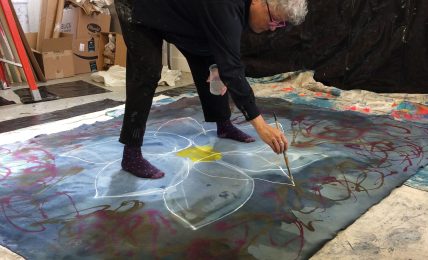 Person with curly white hair painting on large fabric laid on the floor, surrounded by art supplies and boxes.
