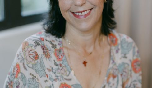 Smiling woman with dark hair and floral blouse sitting indoors near a window.