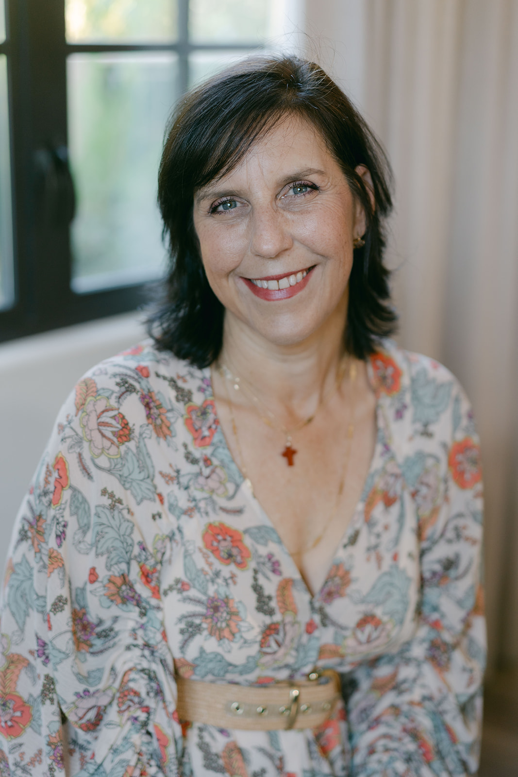 Smiling woman with dark hair and floral blouse sitting indoors near a window.