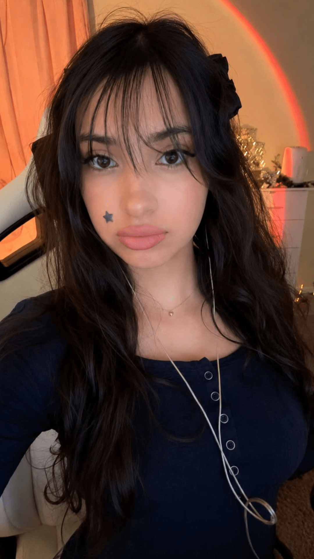 Young woman with long dark hair, black flower hair accessory, star face tattoo, wearing a dark top, indoors.