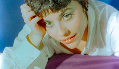 Person with short dark hair lying on a surface, resting their head on their hand, with a colorful object in front.