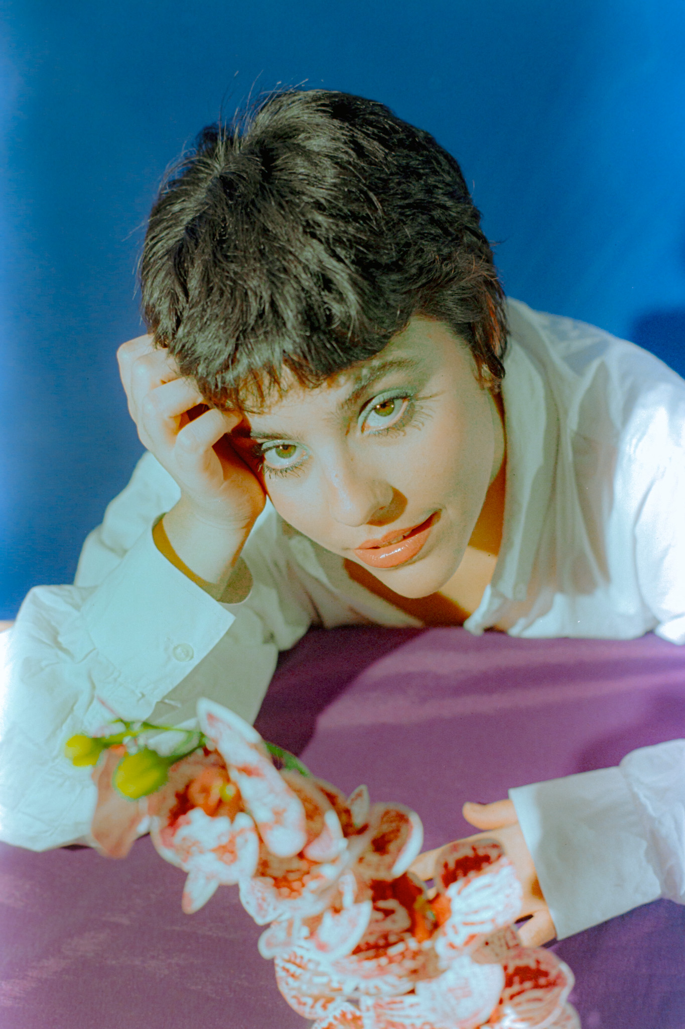 Person with short dark hair lying on a surface, resting their head on their hand, with a colorful object in front.