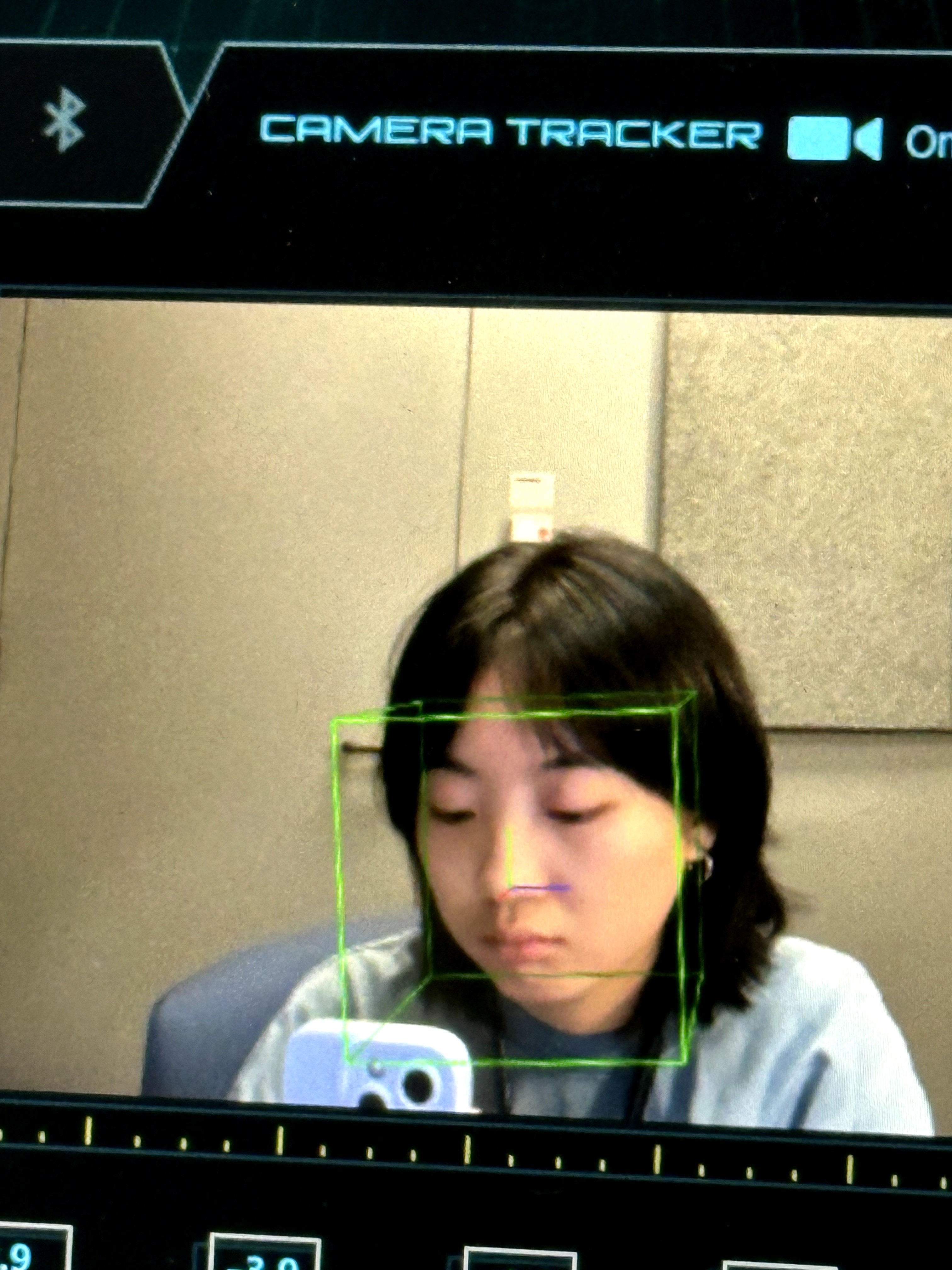 Young woman with dark hair looking down, reflected in a mirror with a green face detection box, in a room with beige walls