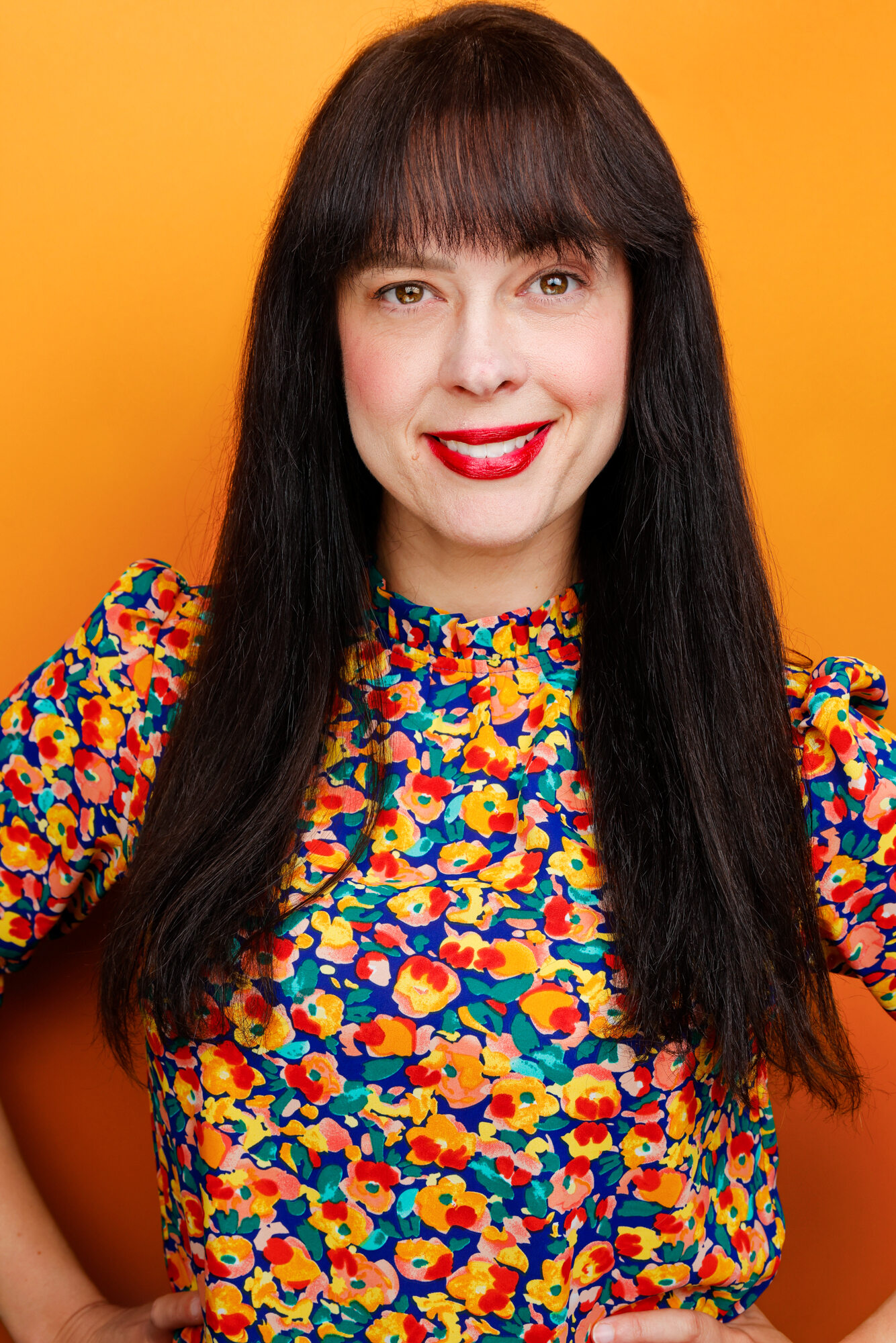 Woman with long dark hair, red lipstick, smiling, wearing a colorful patterned top, standing against an orange background.