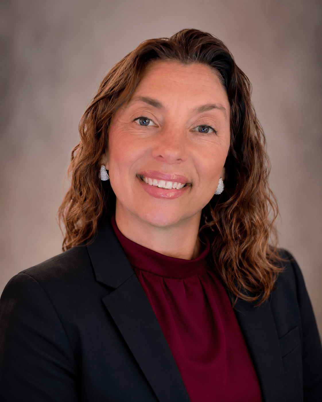 Portrait of a woman with wavy brown hair, smiling, wearing a black blazer and maroon blouse, against a neutral background.