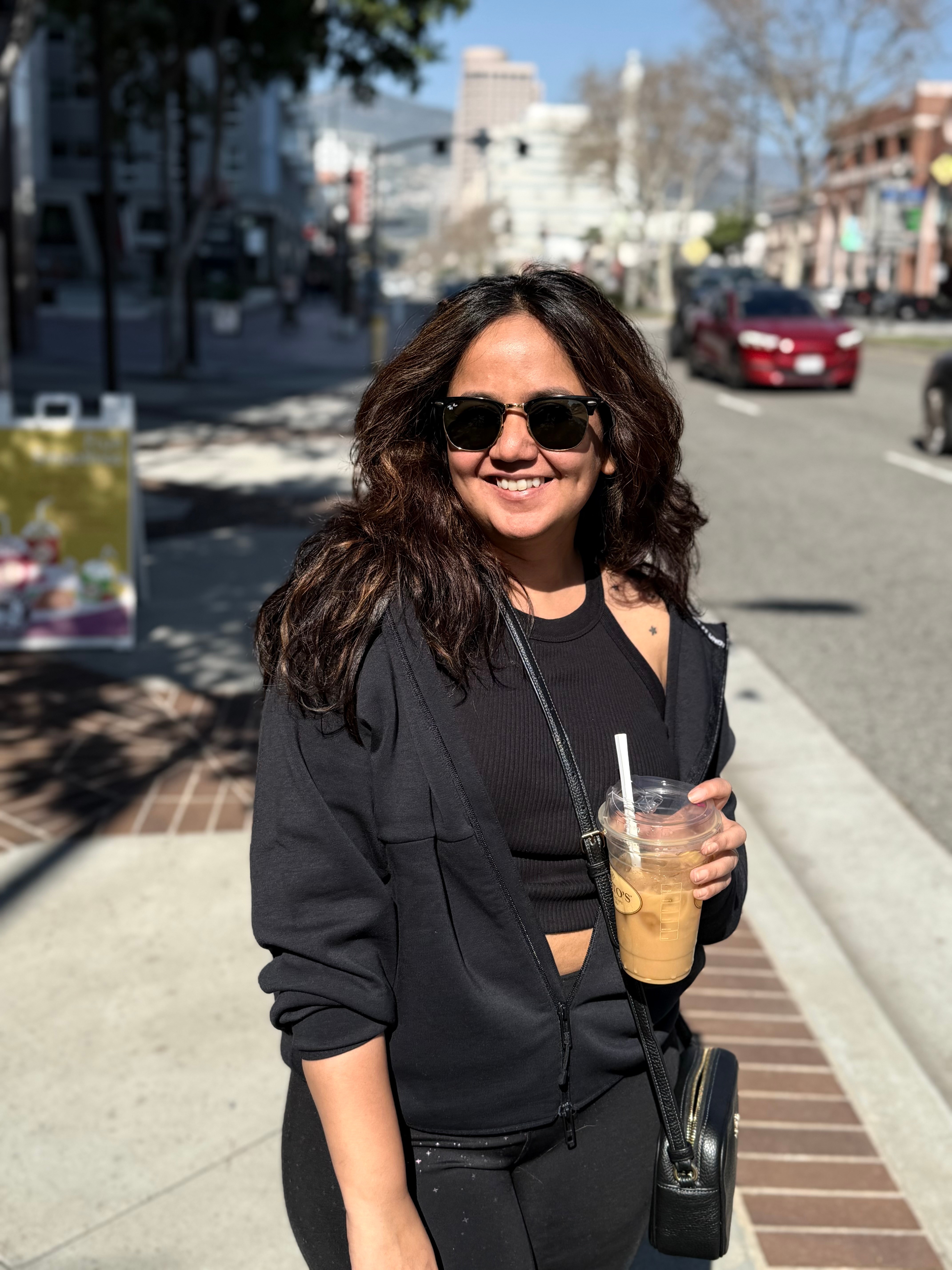 Woman with sunglasses smiling holding a drink on a city sidewalk, sunny day, buildings and cars in background.