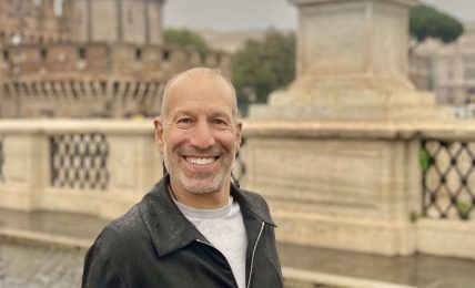 Man smiling outdoors near a fountain with a statue of an angel holding a cross, cloudy sky background.