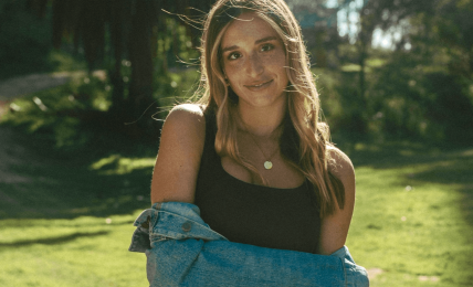 Young woman with long hair standing outdoors in sunlight, wearing a black top and denim jacket, smiling.