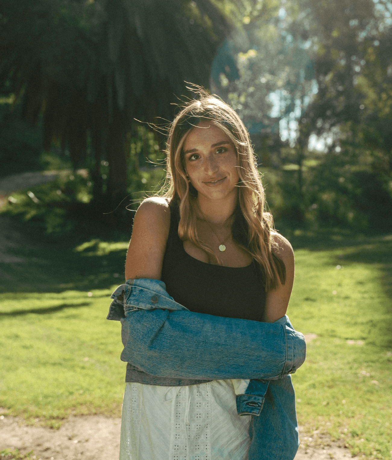 Young woman with long hair standing outdoors in sunlight, wearing a black top and denim jacket, smiling.
