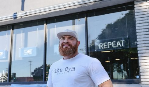 Man with a beard and white cap standing outside gym with glass windows and sign reading 'The Gym'.