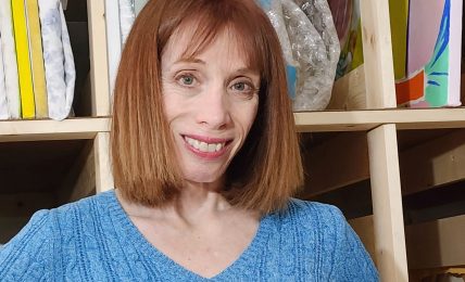 Woman with shoulder-length red hair smiling, wearing a blue sweater, standing in front of shelves with fabric and supplies.