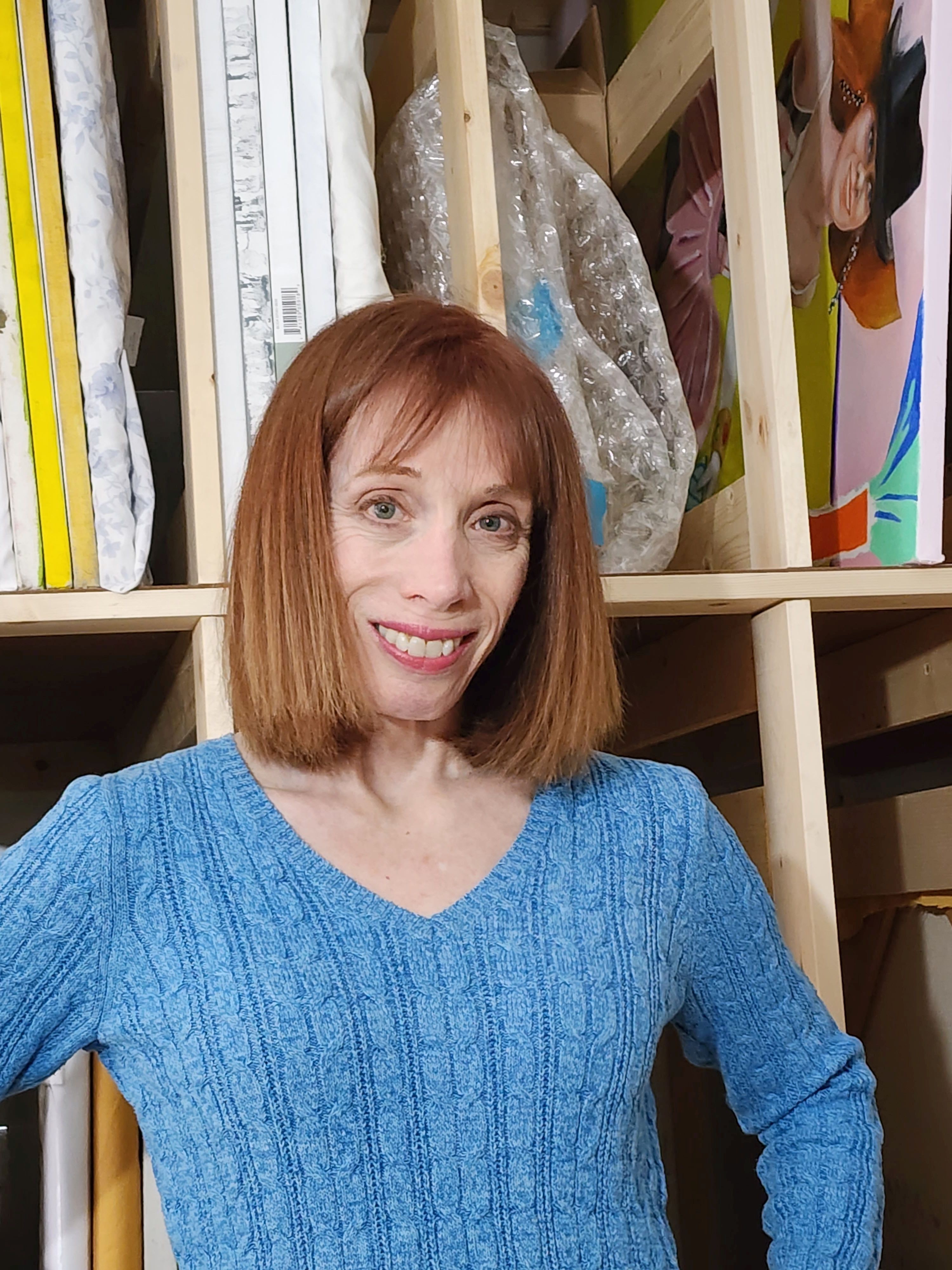 Woman with shoulder-length red hair smiling, wearing a blue sweater, standing in front of shelves with fabric and supplies.
