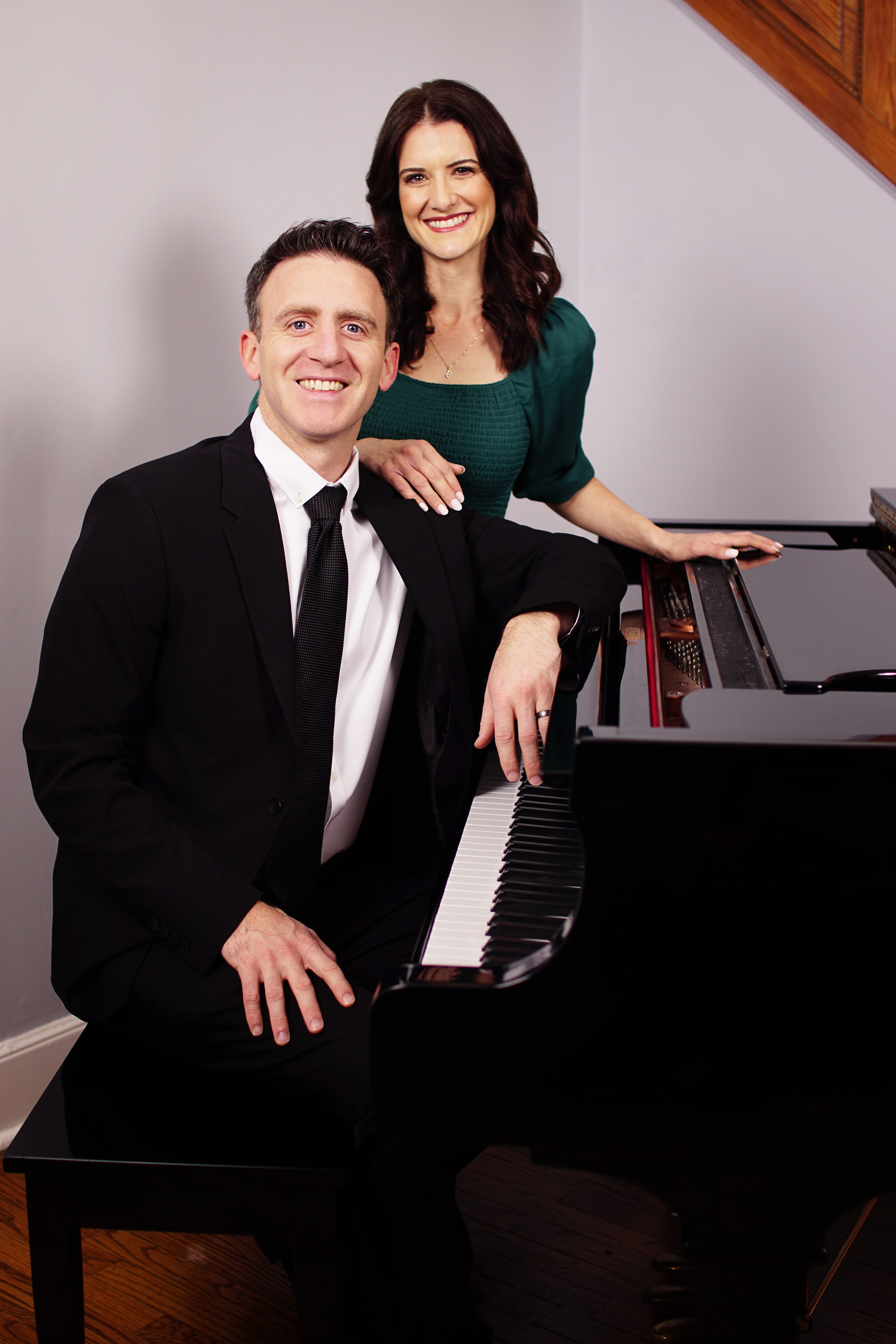 A man in a black suit and a woman in a green top sit at a piano, smiling, in a room with a staircase.