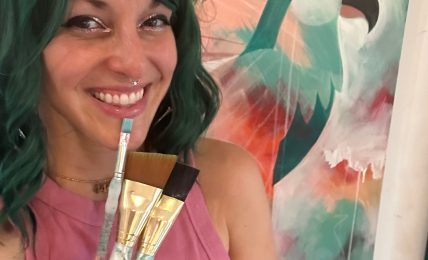 Woman with green hair smiling, holding paintbrushes, in front of colorful abstract background with a bird silhouette.