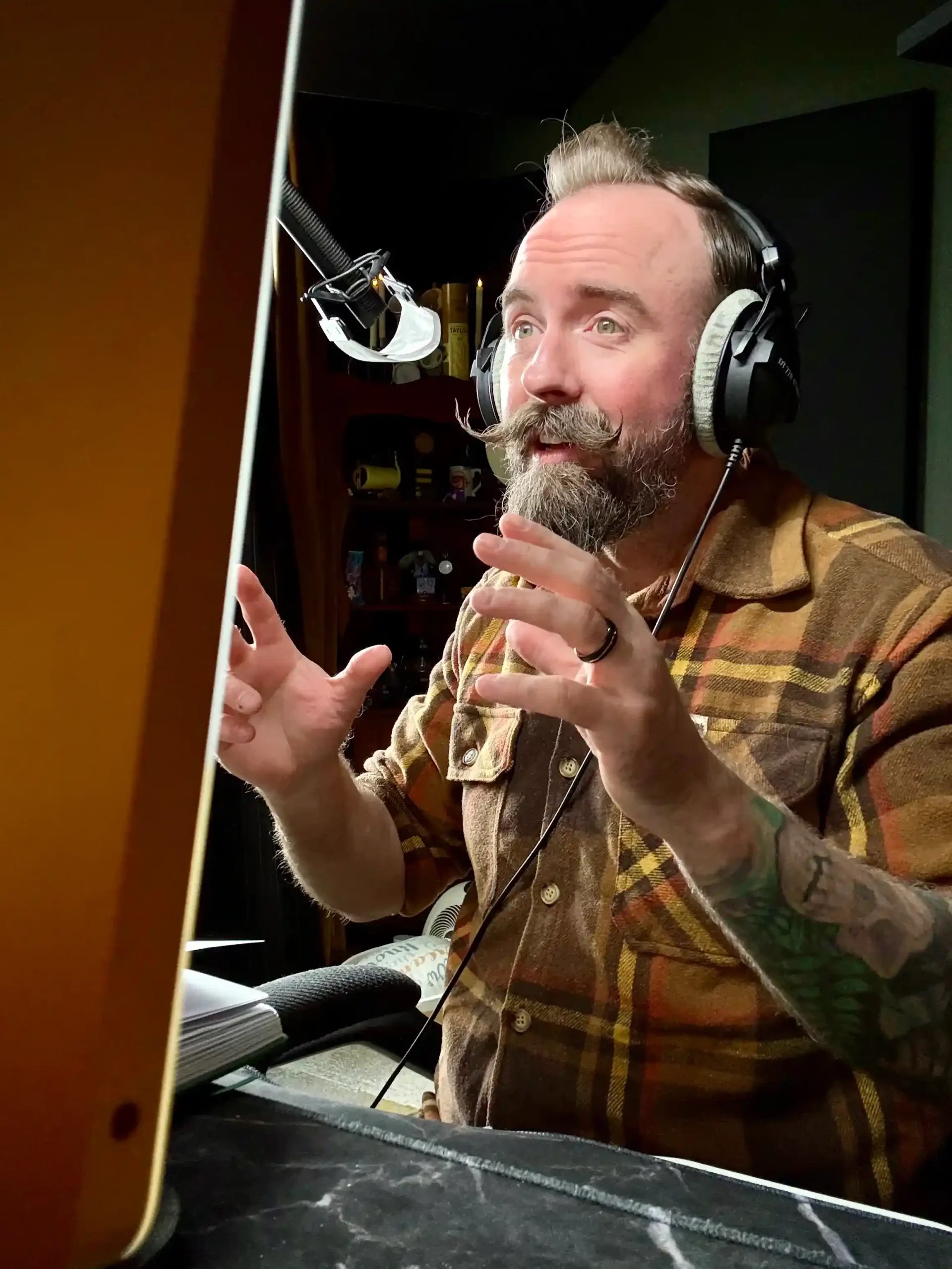 Man with a beard and tattoos wearing headphones speaking into a microphone in a recording studio.