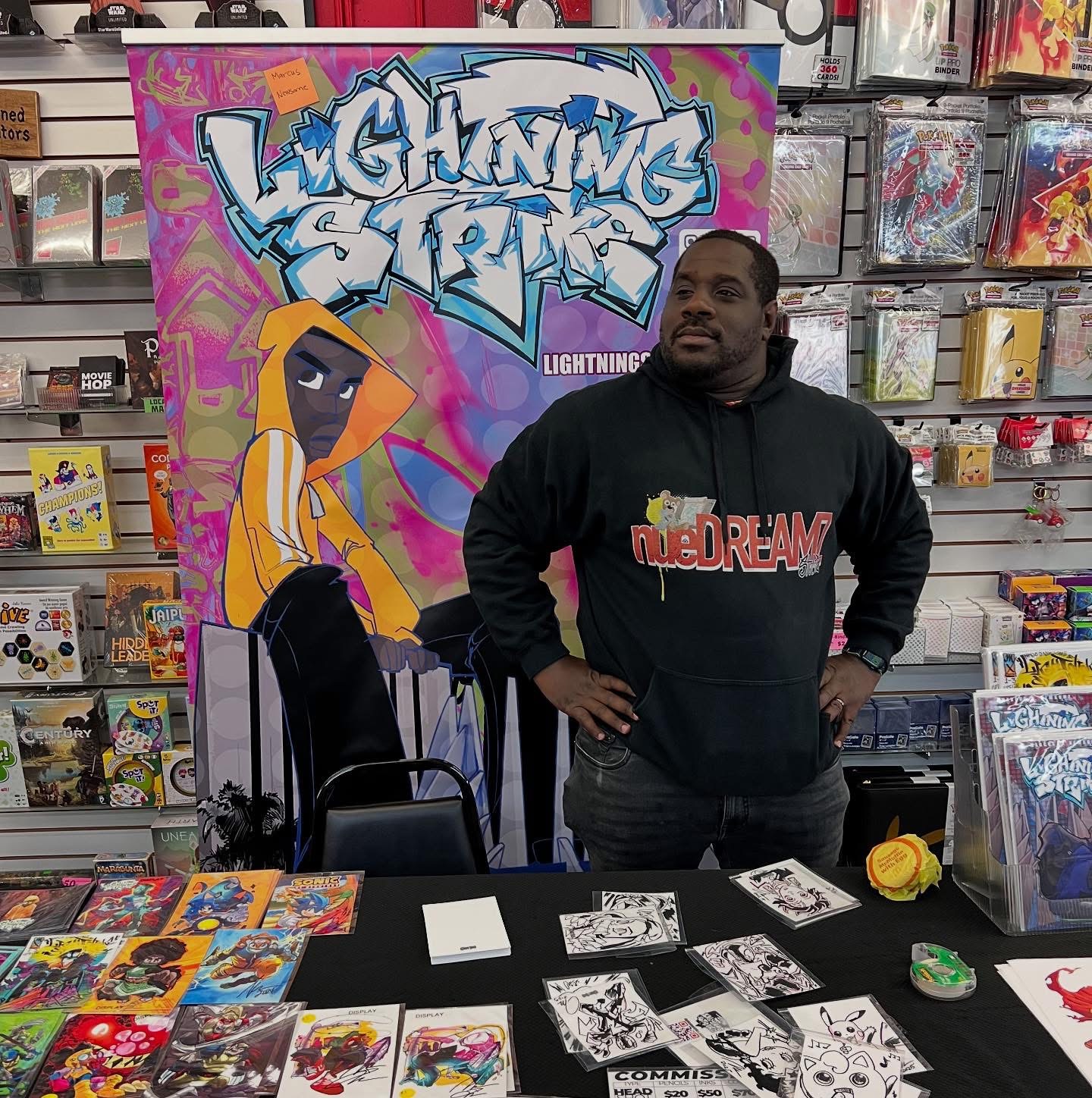 Man standing at a table with comic books, in front of a colorful graffiti-style sign reading 'Lighting Strike'.