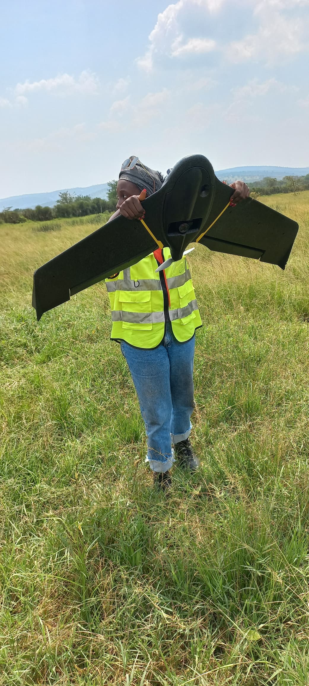 Person in a yellow safety vest holding a large black drone outdoors in a grassy field.