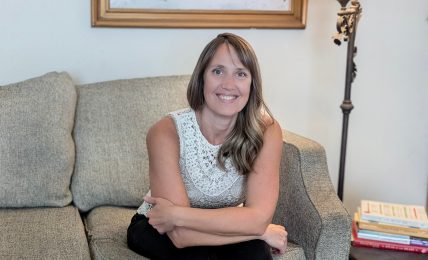 Woman sitting on a sofa in a living room, smiling, with a world map painting and a floor lamp behind her.