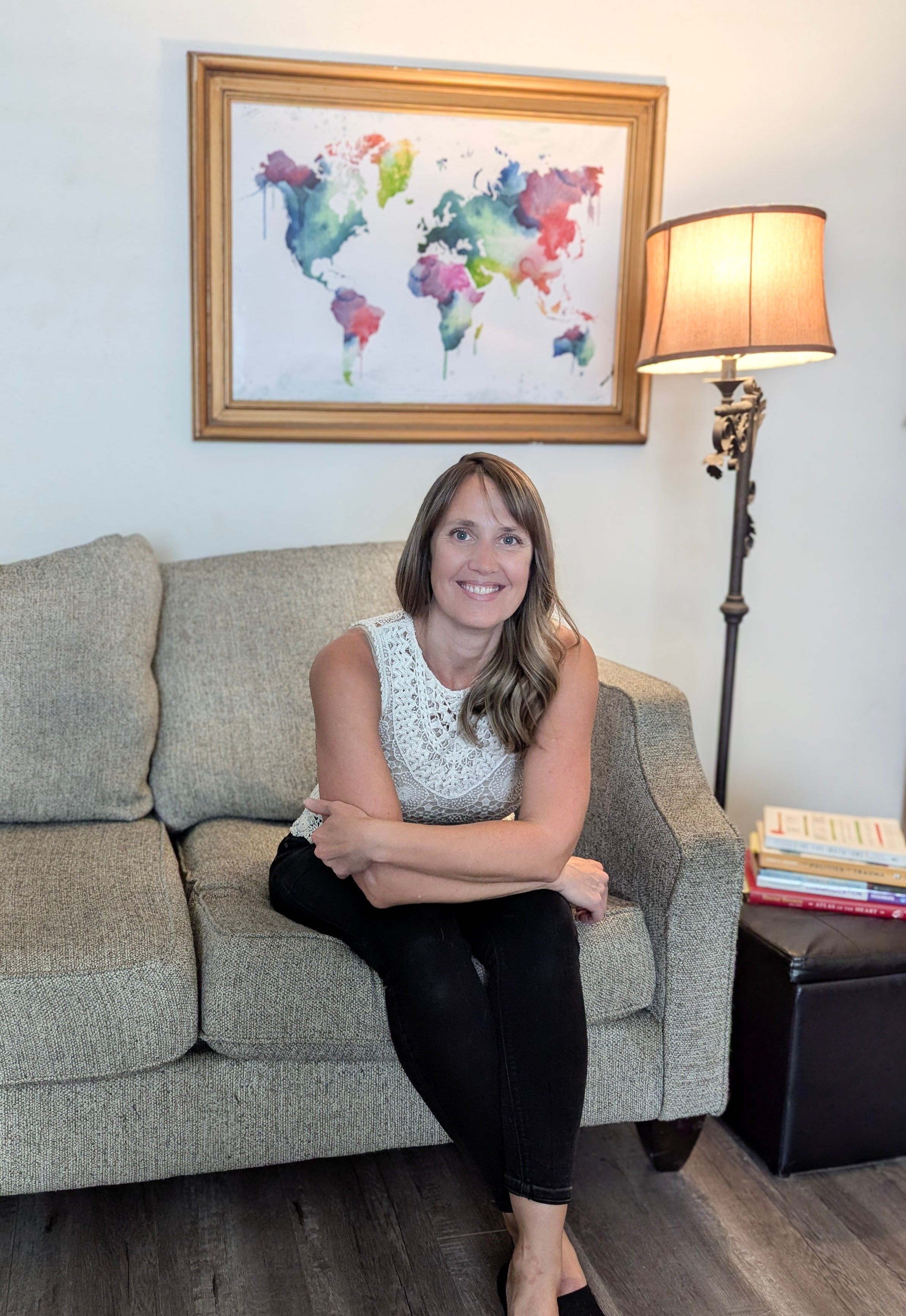 Woman sitting on a sofa in a living room, smiling, with a world map painting and a floor lamp behind her.