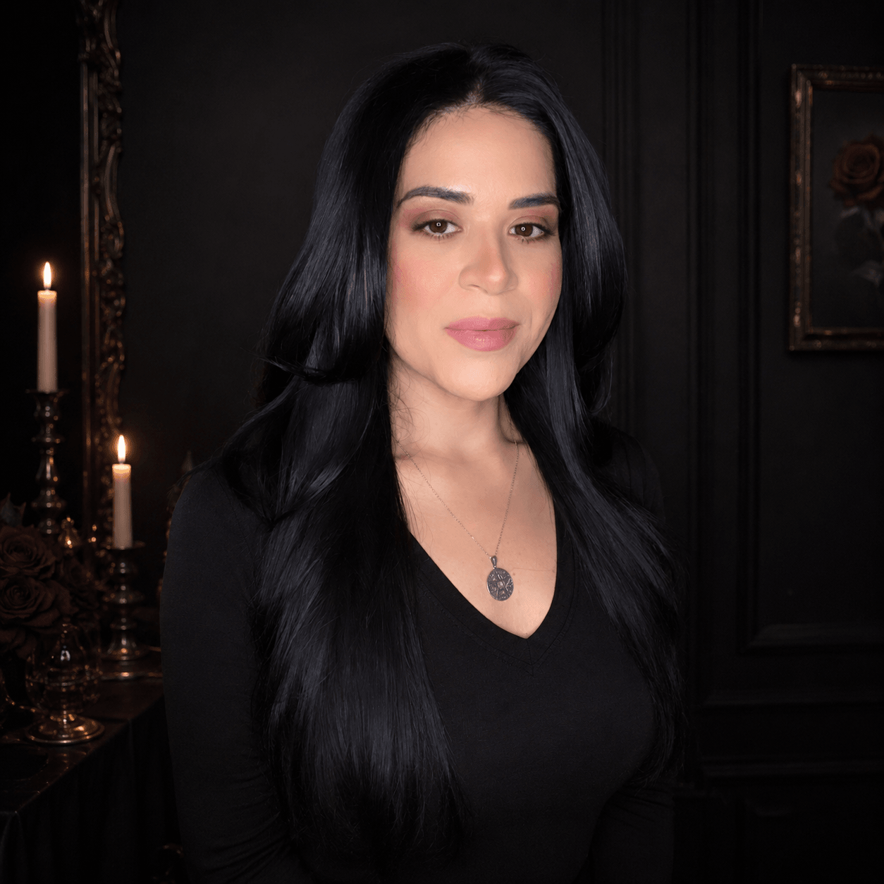 Woman with long black hair and a necklace, standing indoors with dark walls and candles in the background.