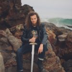 Person with long curly hair holding a sword, sitting on rocks near the ocean, wearing a leather jacket and jeans.
