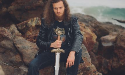 Person with long curly hair holding a sword, sitting on rocks near the ocean, wearing a leather jacket and jeans.