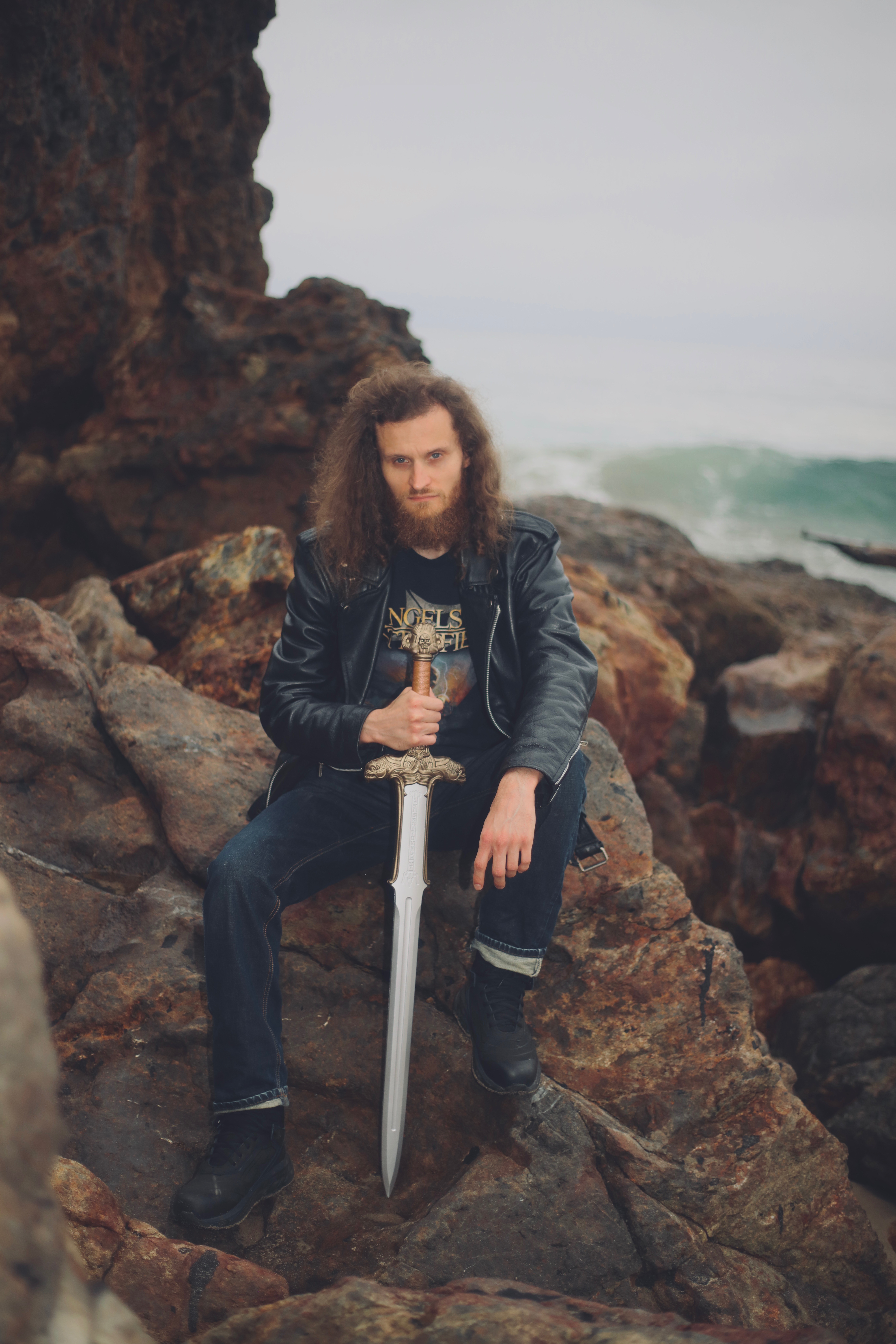 Person with long curly hair holding a sword, sitting on rocks near the ocean, wearing a leather jacket and jeans.