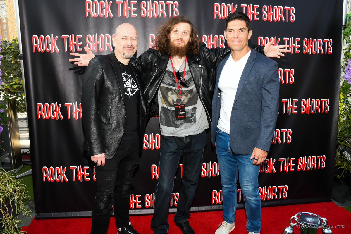 Three men standing together on a red carpet in front of a black backdrop with red text, smiling and posing for the photo.