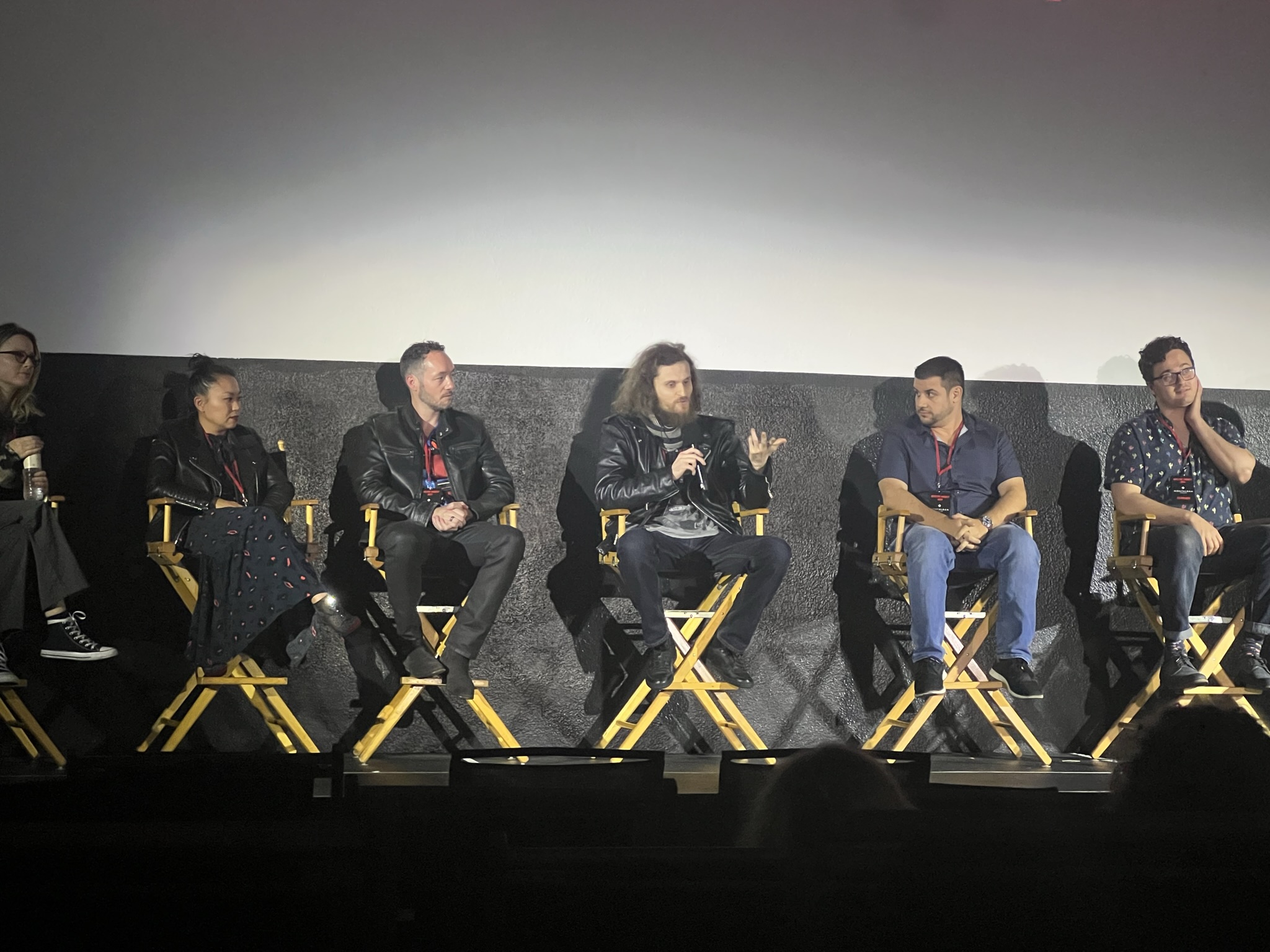 Six people sitting on stage in chairs, engaged in a panel discussion, with a blank screen behind them.